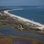 An island with marshes to the west, the ocean to the east. Buildings and a tall water tower next to rocket launch pads to the south with sandy beach.
