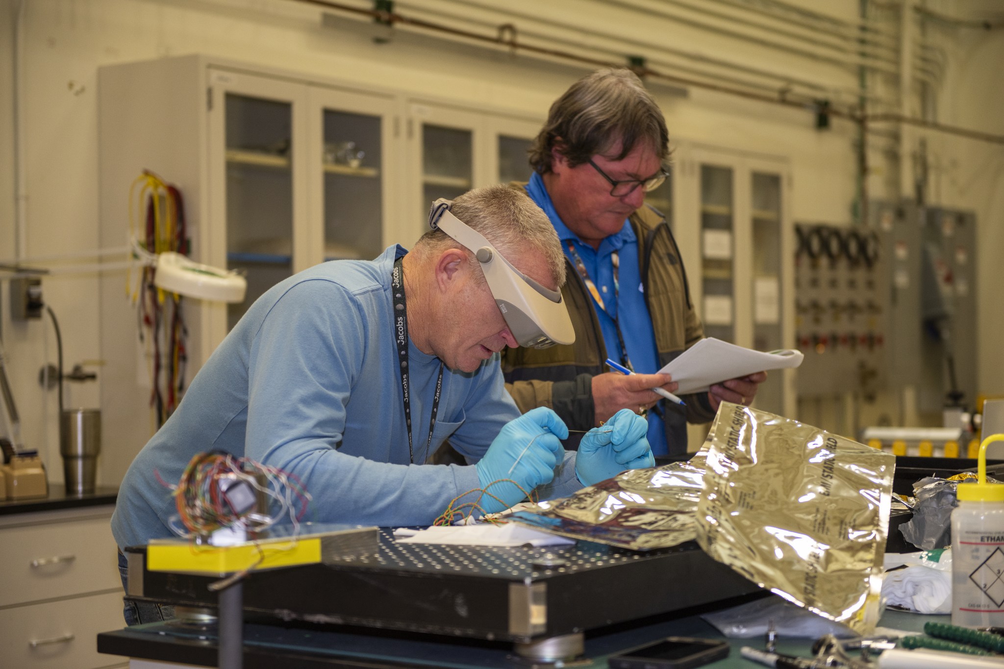 A man wearing blue gloves and a gray visor with a magnifying glass built in it leans against a black table while looking at a specimen. Next to him, a second man wearing black glasses is standing while holding a pen and a stapled stack of papers.