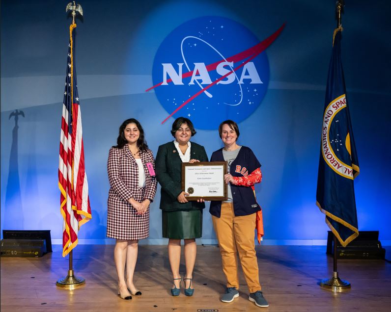An Iranian woman with short hair wearing a green and blue skirt suit accepts a framed recognition from a woman with short hair and wearing a baseball jersey on a stage with an American flag, NASA flag, and NASA meatball.