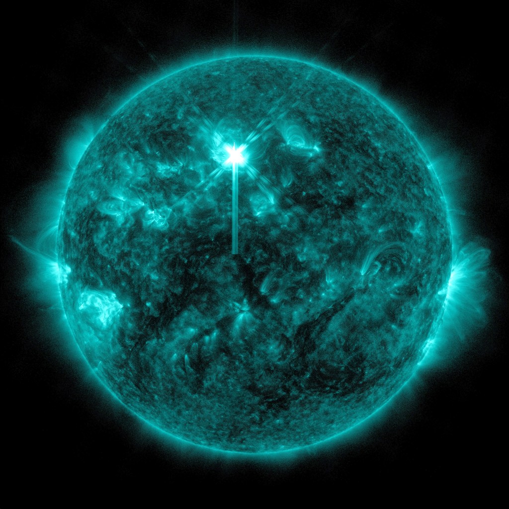 The Sun, colorized in teal. There are many bright active areas on the star, including on the bottom left and bottom right areas. Toward the upper middle of the Sun, there is a bright flash, creating an "X" shape.