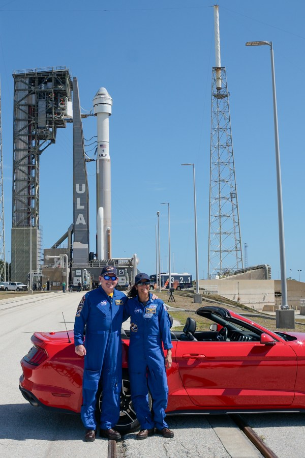 NASA astronauts Butch Wilmore and Suni Williams stand in front of a red convertible with the ULA Atlas V rocket and Boeing Starliner spacecraft on the launch pad in the background