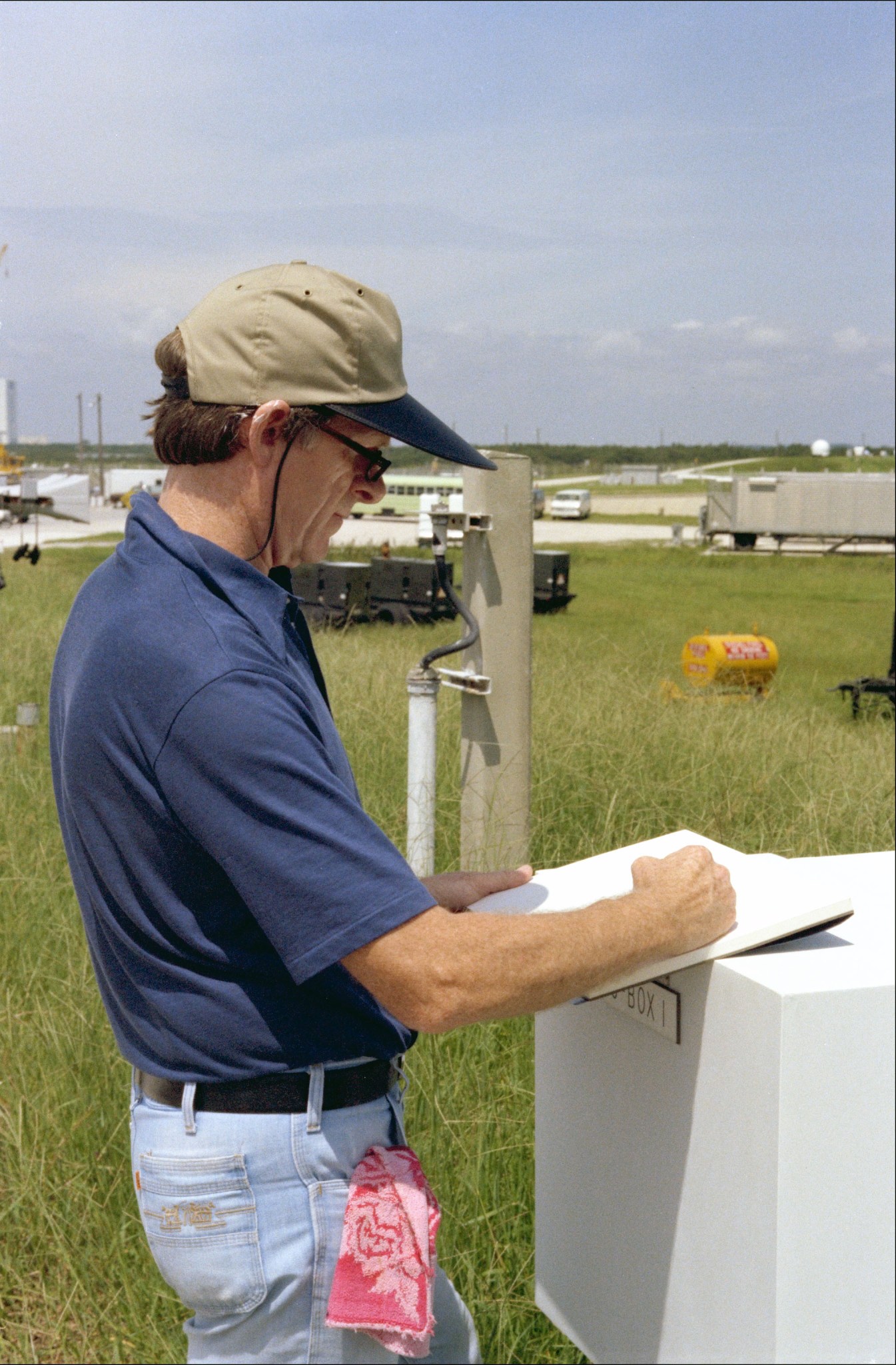 James Dean stands in the grass at the launchpad at NASA's Kennedy Space Center and draws on a sketchpad while using a white box as a makeshift easel. He is wearing a dark blue polo shirt, blue jeans, sunglasses, and a beige baseball cap with a dark rim. A red patterned fabric is sticking out of his right-hand pocket of his jeans.