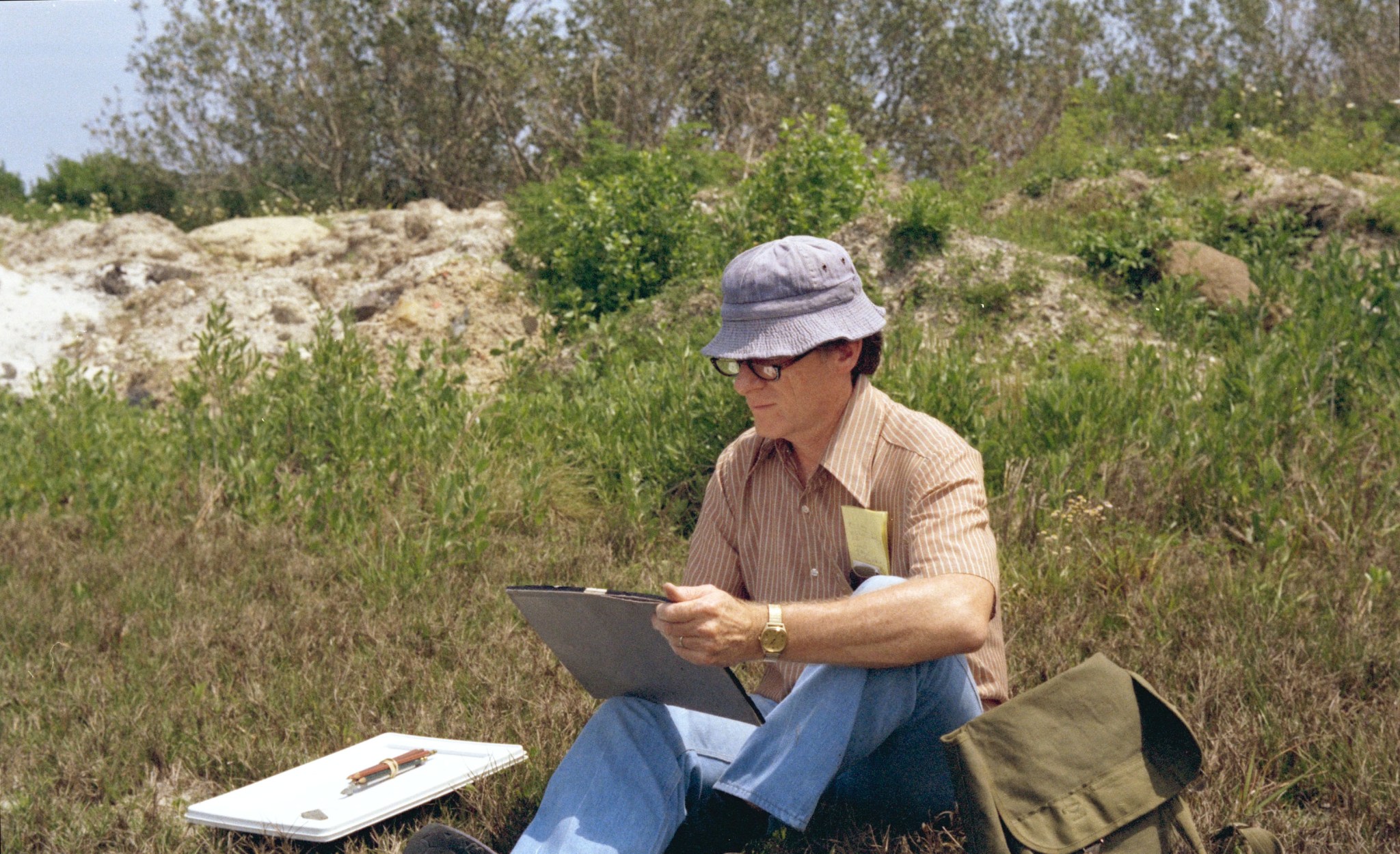 James Dean sits on the grass, holding a sketch pad and looking at it. He is surrounded by artist materials on his left and a green bag on his right. He is wearing a striped orange shirt, blue jeans, a gray bucket hat, and dark glasses.