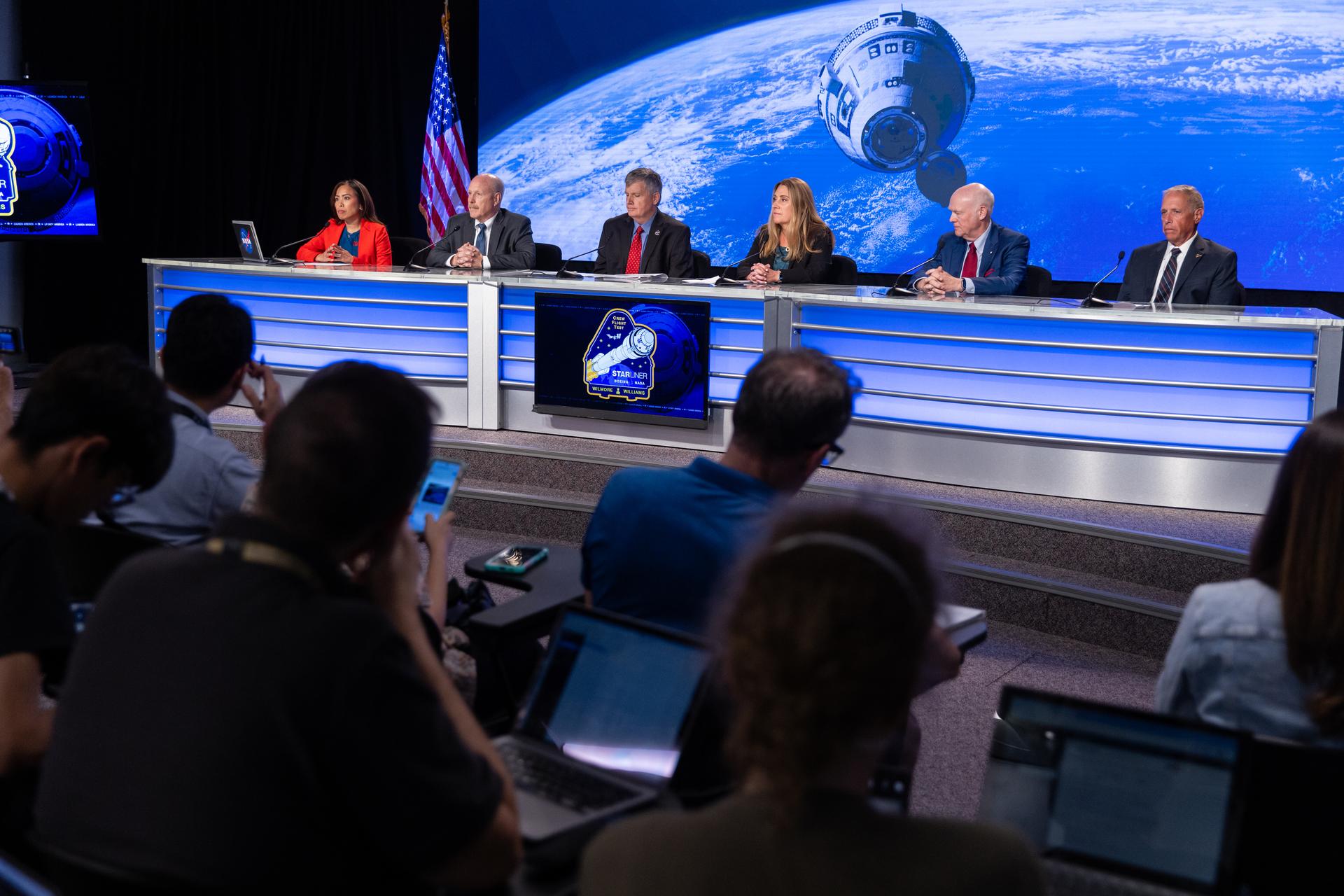 Six people sit at a news desk with a large screen behind them with an image of a spacecraft in front of the Earth.