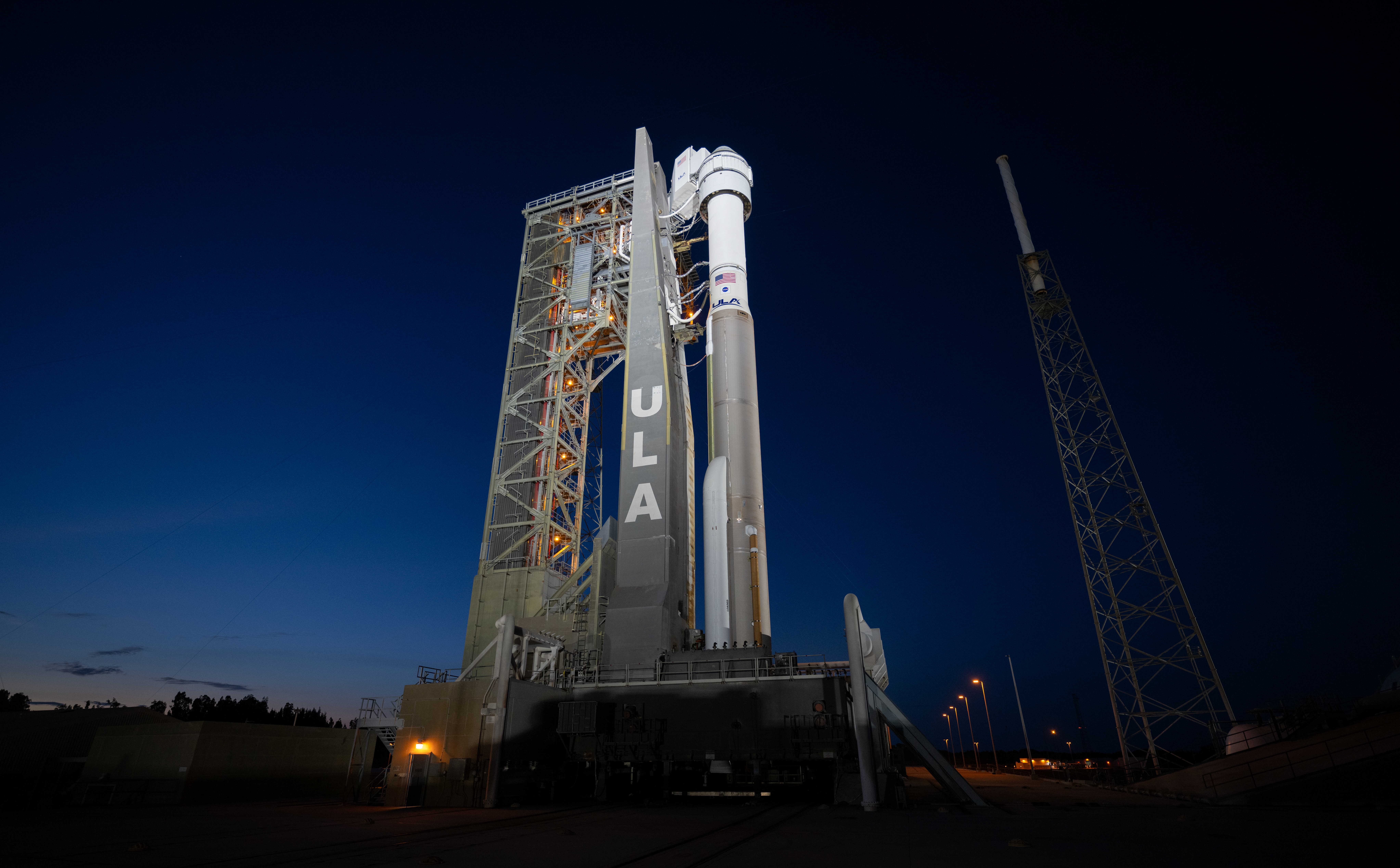 A rocket with a spacecraft atop stand in the center of the image illuminated by spotlights against a dark sunset sky.