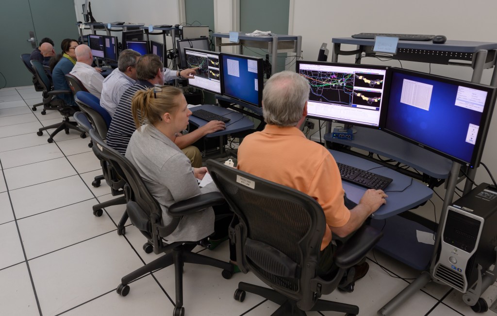 A row of eight simulation participants seated workstations. The MFCR user interface is displayed on their screens.