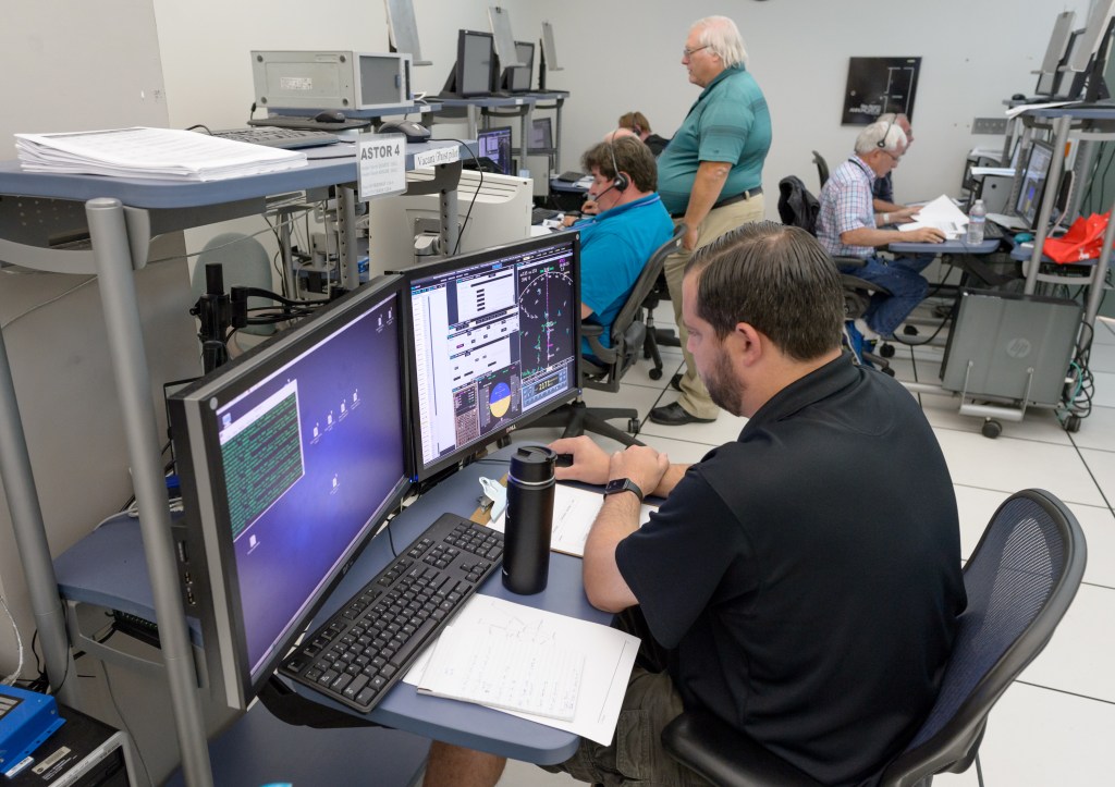Six simulation participants seated at pseudo-pilot stations for the UAM HITL simulation. A NASA researcher stands to observe one of the station displays.