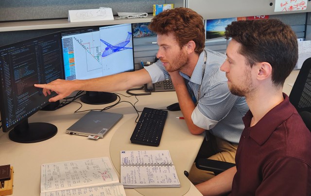 两名男子在NASA办公室的办公桌旁工作，其中一人指着显示器上显示的一些Aviary计算机代码。未来飞机设计的图片出现在邻近的显示器上。