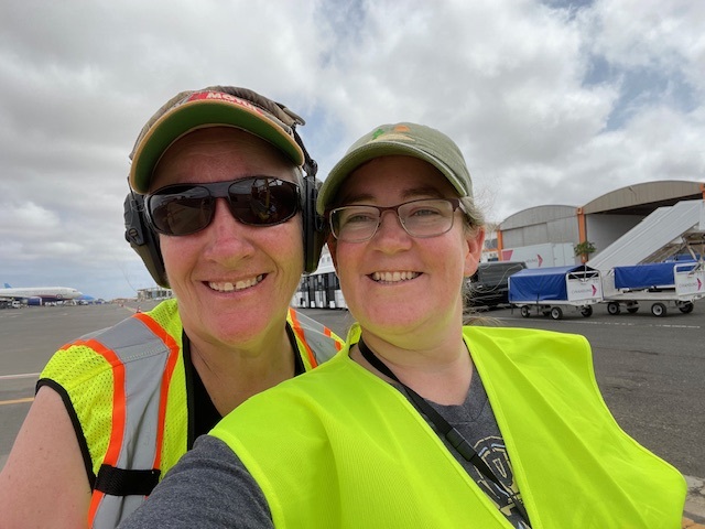 Two women smile close-up at the camera. Both women are wearing bright reflective vests and caps; the woman on the left is wearing sunglasses.