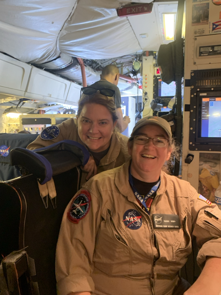 Two women pose and smile at the camera. Both are wearing tan flysuits and sitting at the mission control desk in the interior of an aircraft.