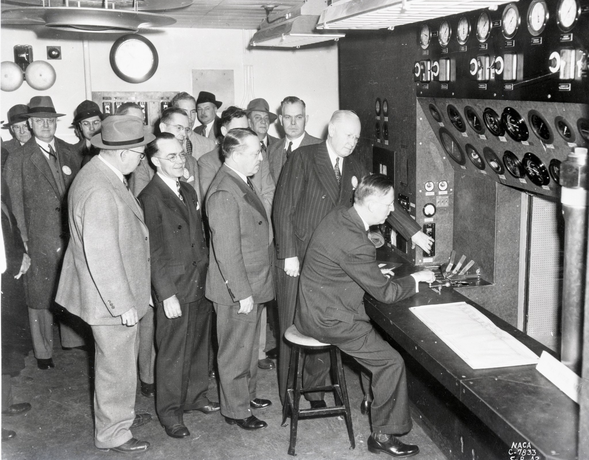 A black-and-white image of a large group of men wearing suits in a control room. One NACA leader presses a button, and another spins a crank.