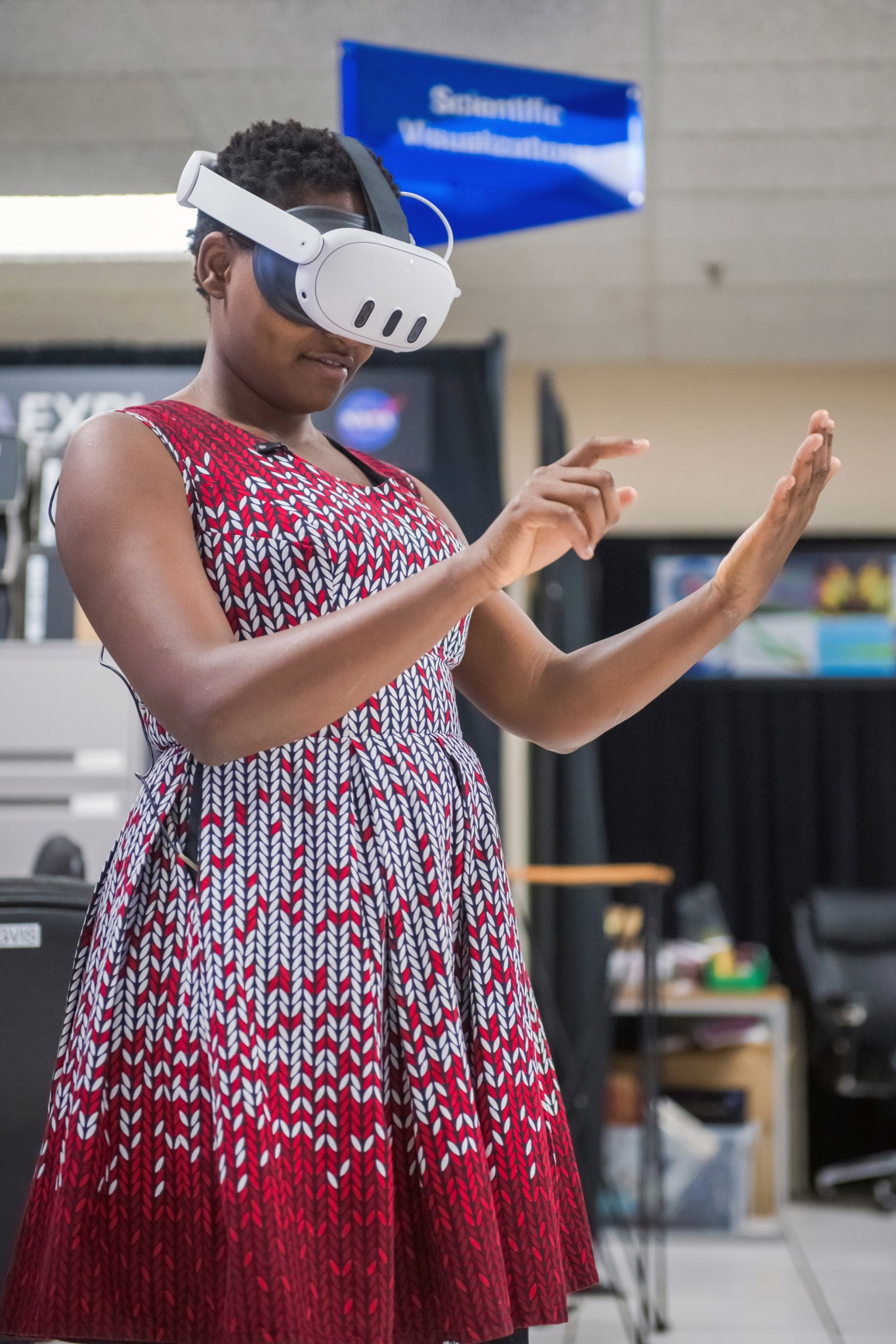 Nelly Cheboi concentrates while testing a virtual reality demonstration. She wears a virtual reality headset and a red and white dress. Her hands appear as if she is pressing buttons on an invisible tablet.
