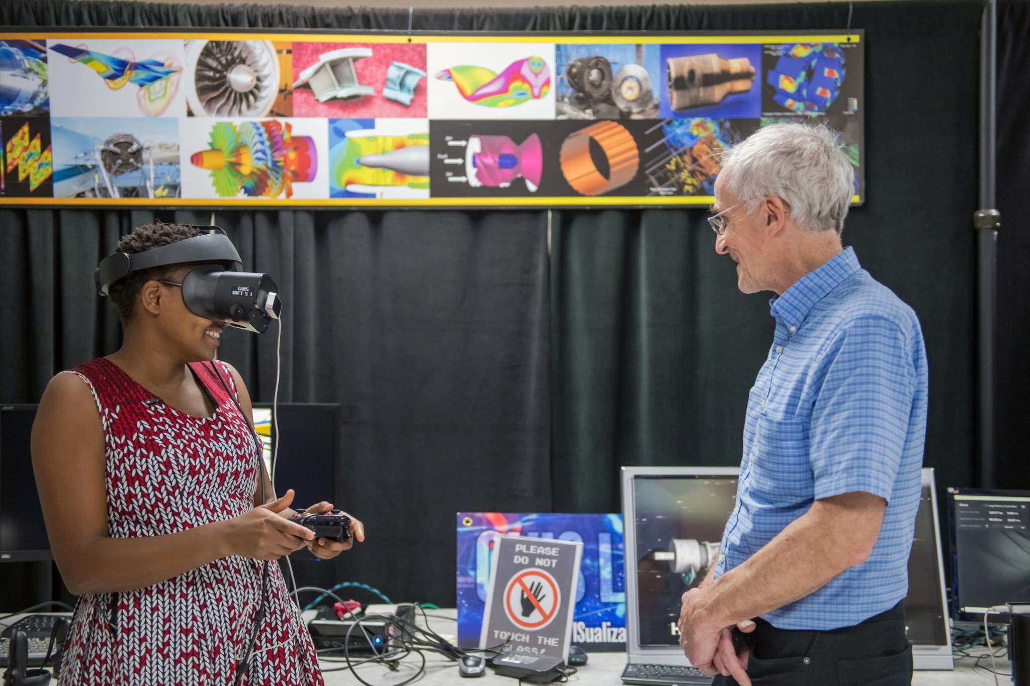 Nelly Cheboi enjoys a virtual reality demonstration. She smiles as she wears a VR headset and holds a remote controller. She is wearing a red and white dress. Herb Schilling, computer scientist, smiles as he watches her.