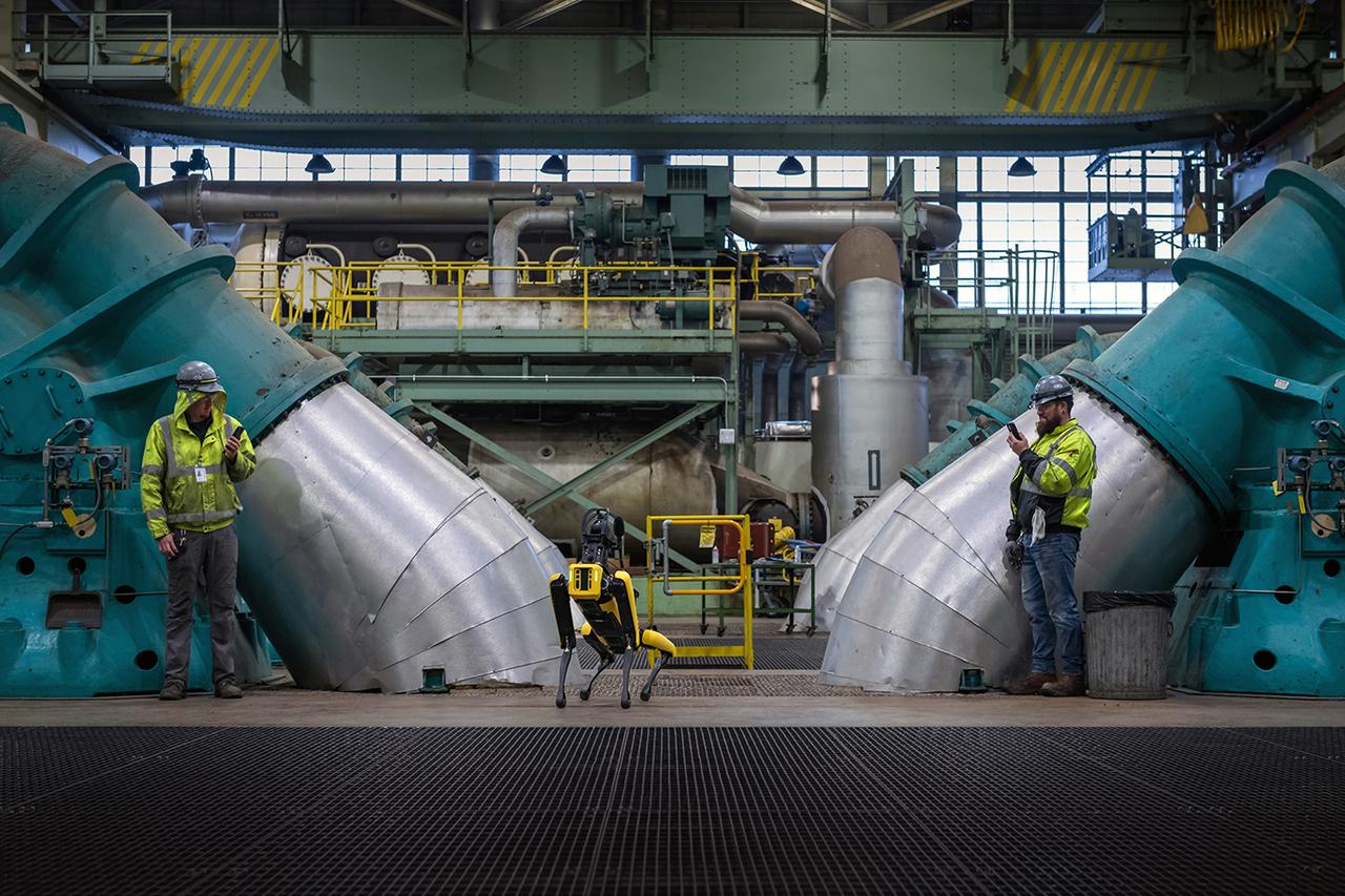 Looking inside NASA Glenn’s Central Air Equipment building showing two large compressors with an employee in front of each compressor. A robotic dog stands in the center and awaits commands.