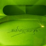 A NASA insignia, also known as the meatball, is being 3D printed and appears to be green in its printing chamber with a bright laser flash in the upper right-hand corner.