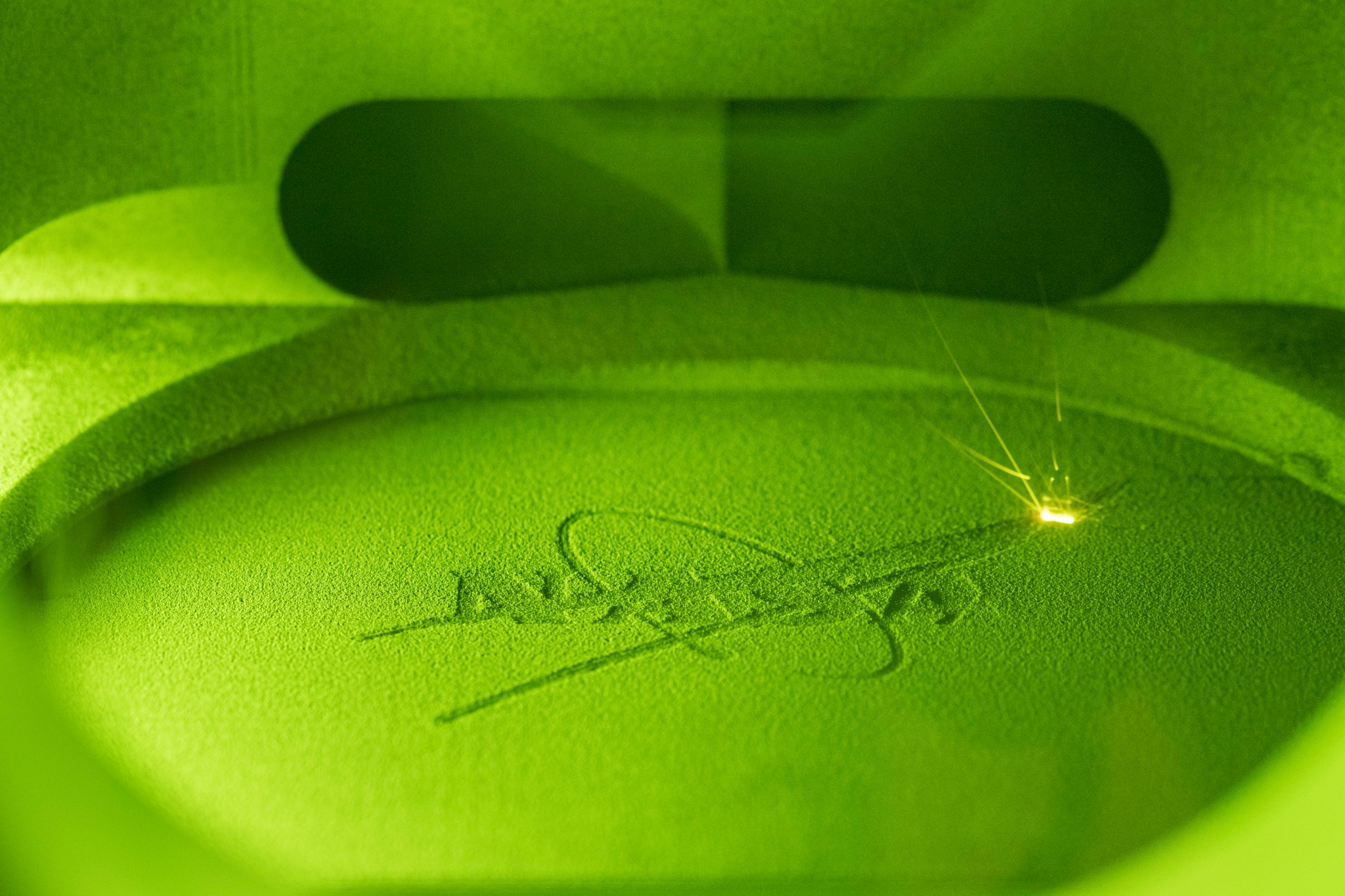 A NASA insignia, also known as the meatball, is being 3D printed and appears to be green in its printing chamber with a bright laser flash in the upper right-hand corner.