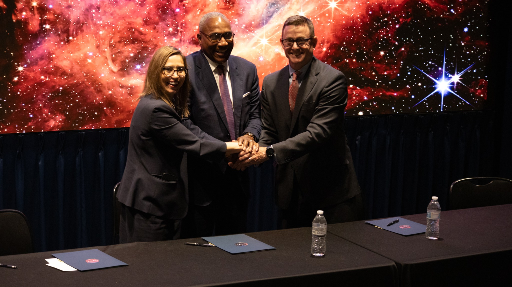 One woman and two men smile with their hands together in a team huddle in front of a large display of an orange-red galaxy. Three folders containing a memorandum of understanding are on a black table with the NASA meatball in front of them.