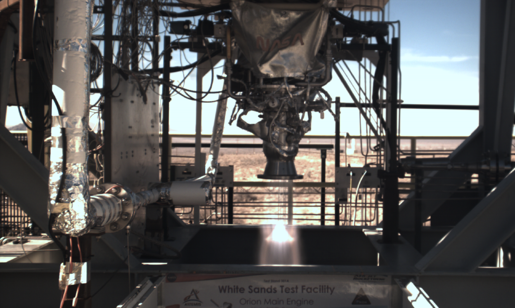 A rocket engine mounted to a metal test stand is firing.