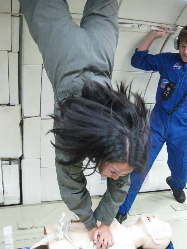 A woman floating upside down in microgravity wearing a light brown flight suit pushes her hands against a