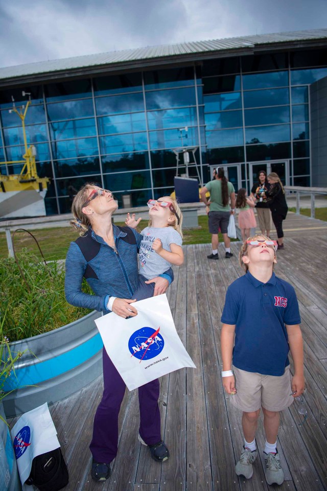 A family of three, all wearing eclipse glasses, stare at the sky while standing outside of INFINITY Science Center