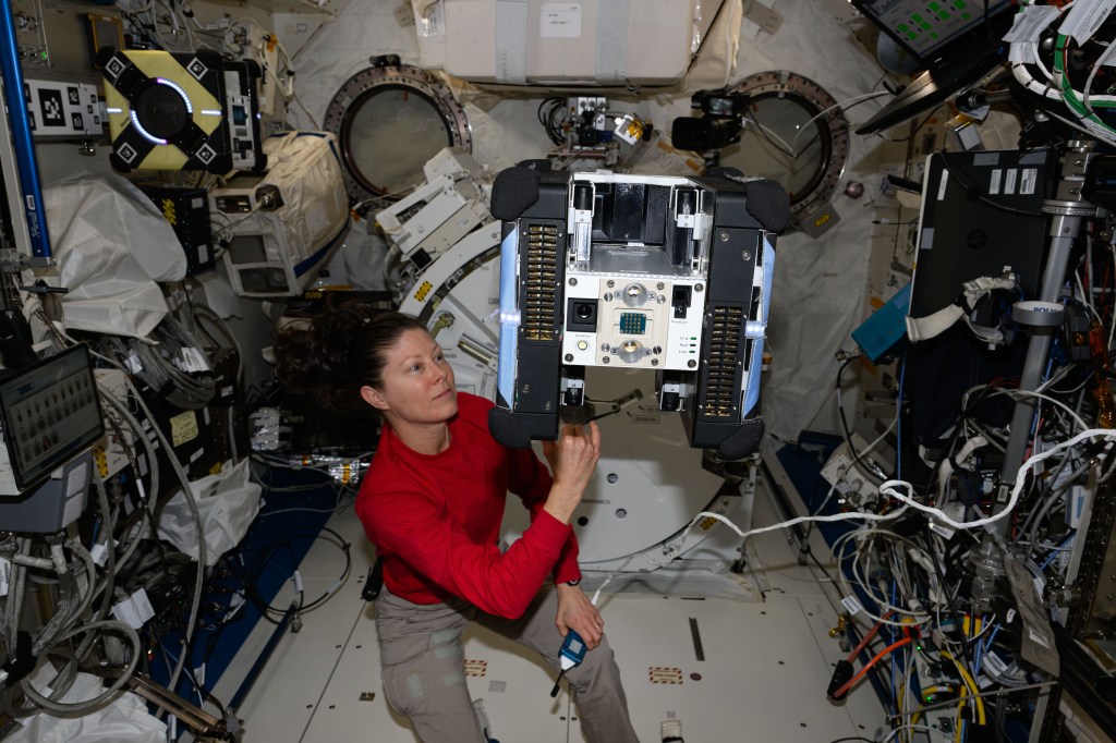 NASA astronaut Tracy C. Dyson, wearing a long-sleeved red shirt and khaki pants, is touching an Astrobee floating just above her left shoulder with her right hand and holding a microphone in her left hand. The robot has a blue panel on each side, black panels inside those, and a white and silver center with two camera lenses and multiple buttons on the front of it.