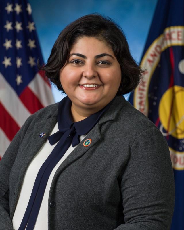 Official headshot of an Iranian woman with short hair, wearing a gray blazer over a blue and white blouse, with an American flag and a NASA flag in the background.