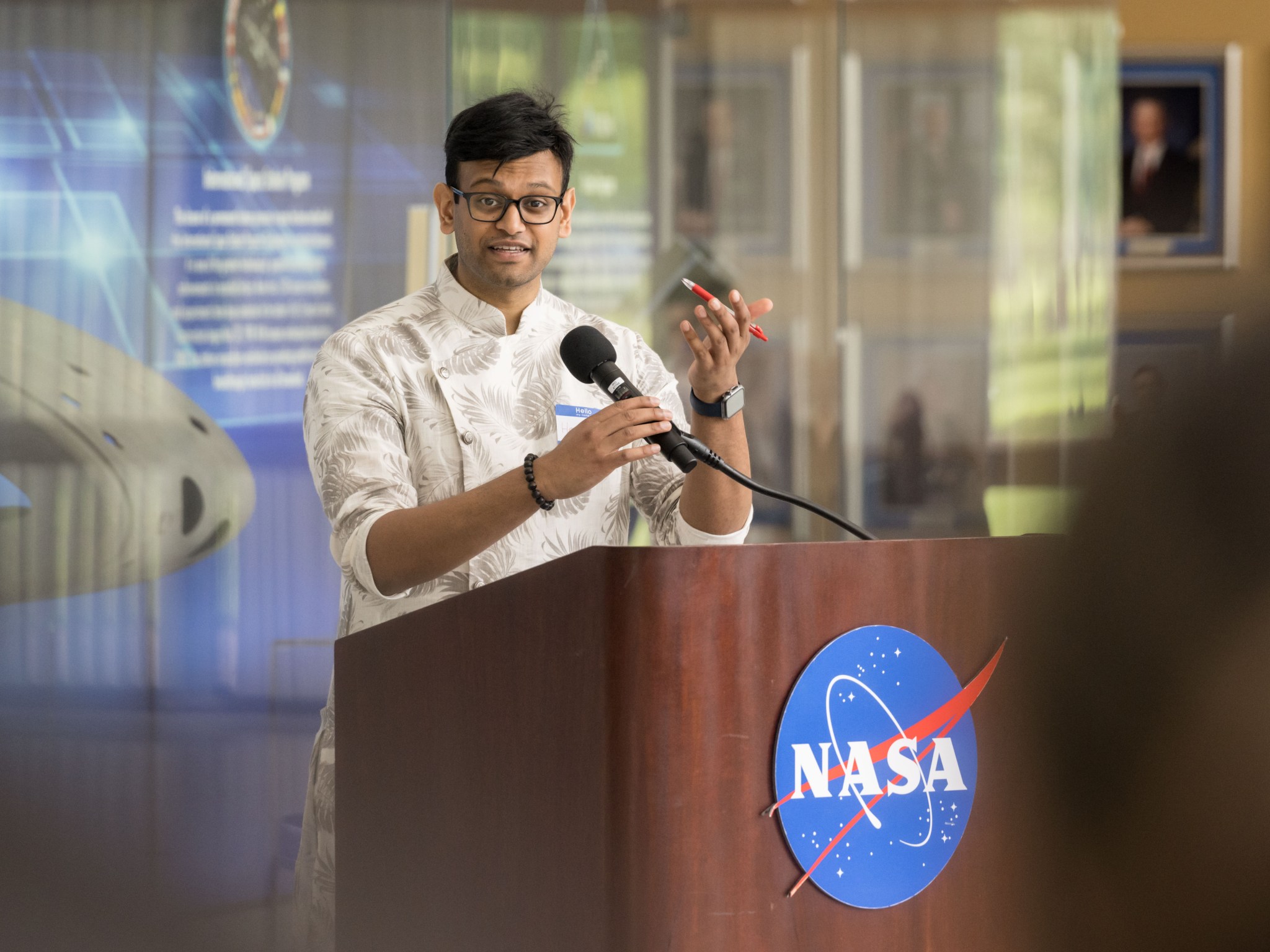 An Indian man wearing glasses and traditional attire speaks at a podium with the NASA meatball on the front.