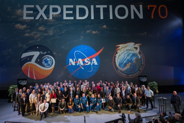 A large group photo taken indoors. The background features three large insignias: one for the International Space Station, the NASA logo in the center, and a mission patch on the right.