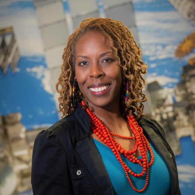 A woman wears a blue top, black button down, and orange necklace in front of a blue sky background with a sketch of the International Space Station.