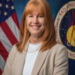 A woman with red hair wearing a white shirt and Khaki blazer poses in front of the United States and NASA flags.