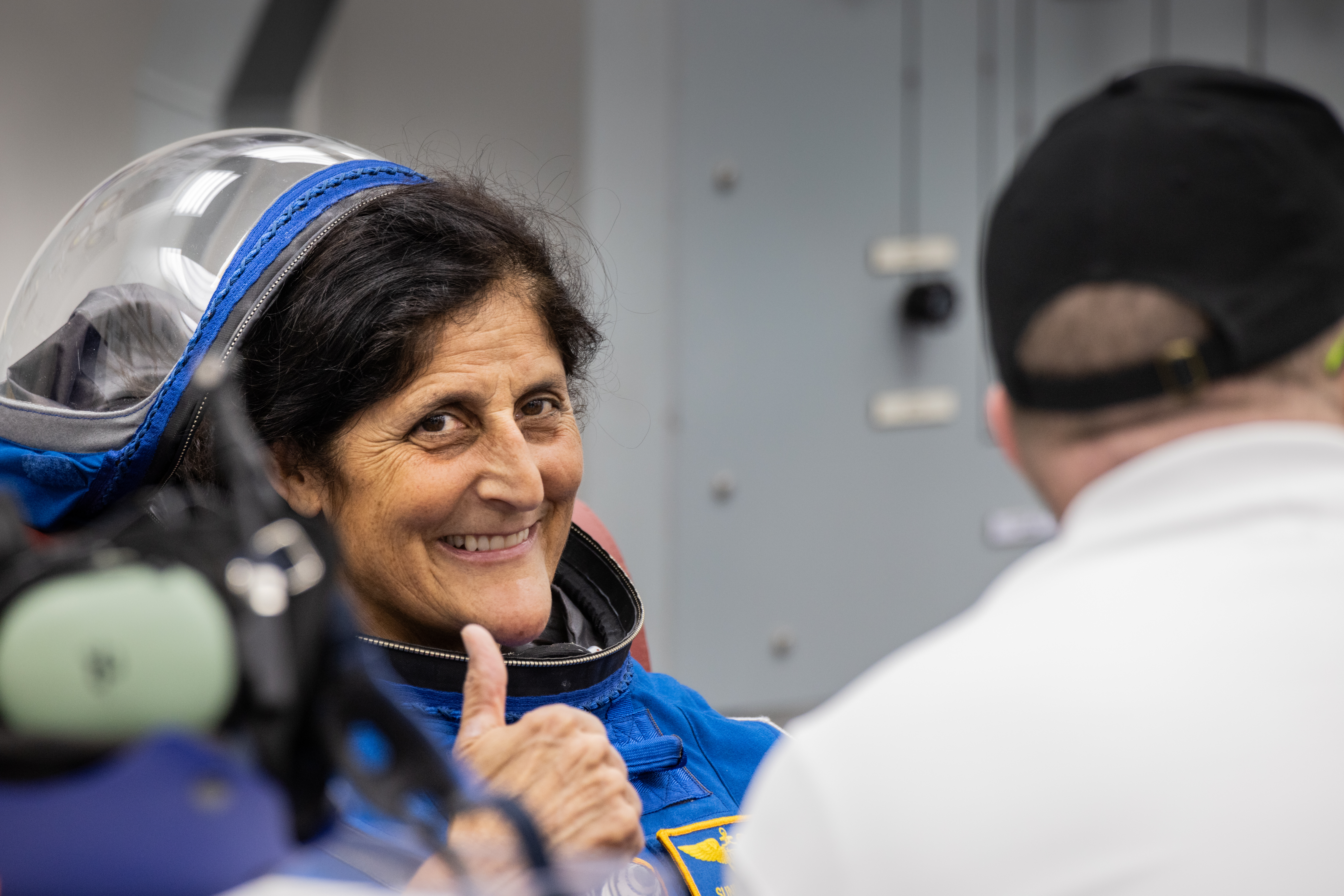 Astronaut Suni Williams (left), an Indian- and Slovene-American woman, smiles and gives a thumbs up to the camera. She wears a blue Boeing spacesuit.