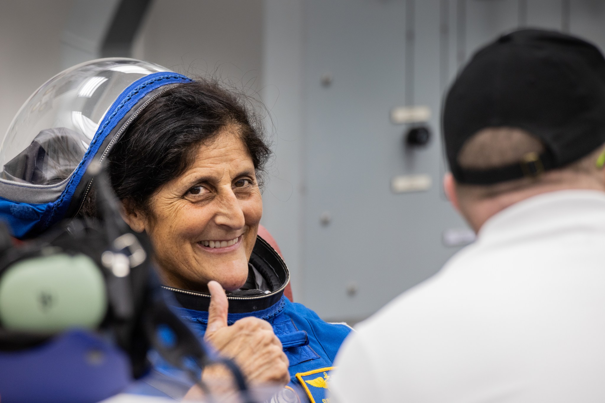 Astronaut Suni Williams (left), an Indian-American woman, smiles and gives a thumbs up to the camera. She wears a blue Boeing spacesuit.