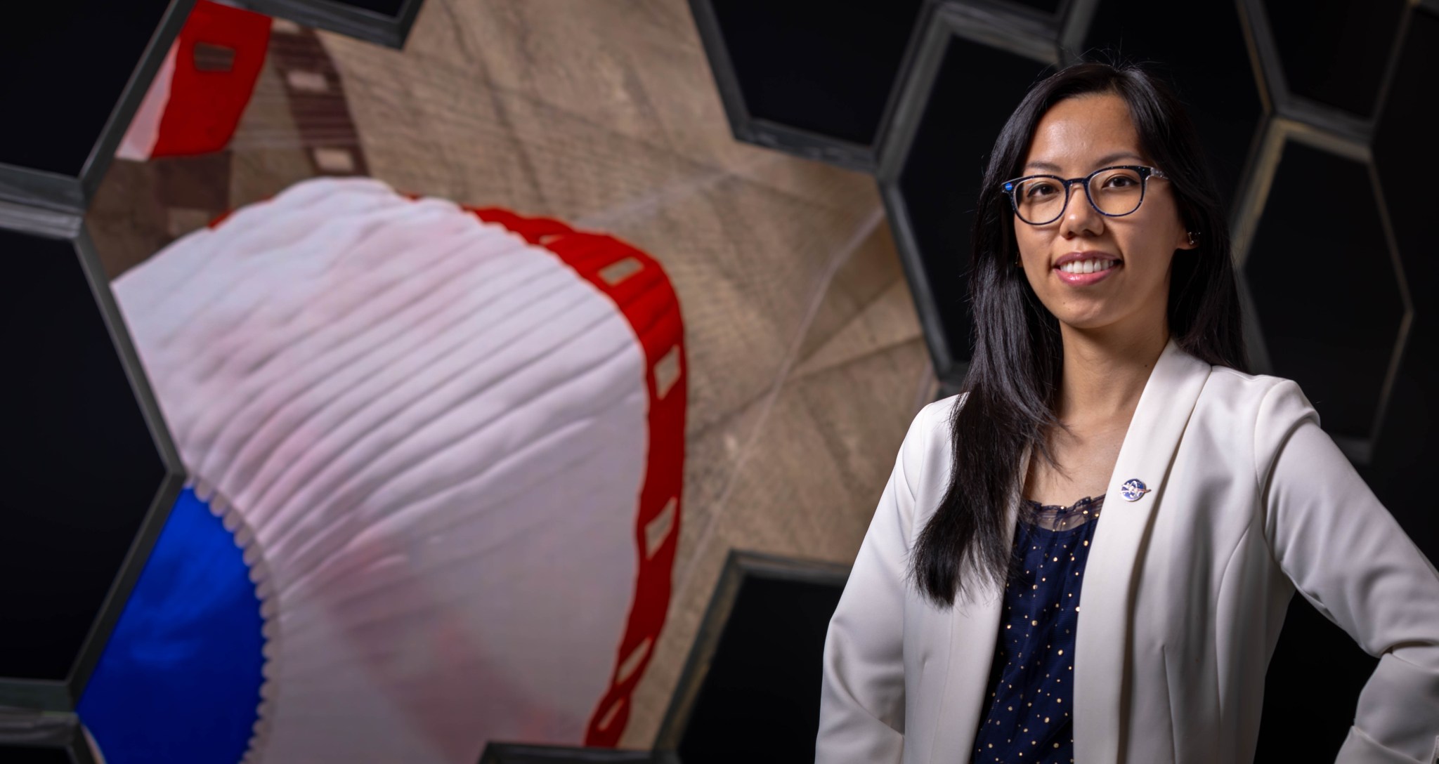 Parachute Engineer Anh Nguyen poses with her hands on her hips in a white blazer, smiling at the camera wearing black rimmed glasses.