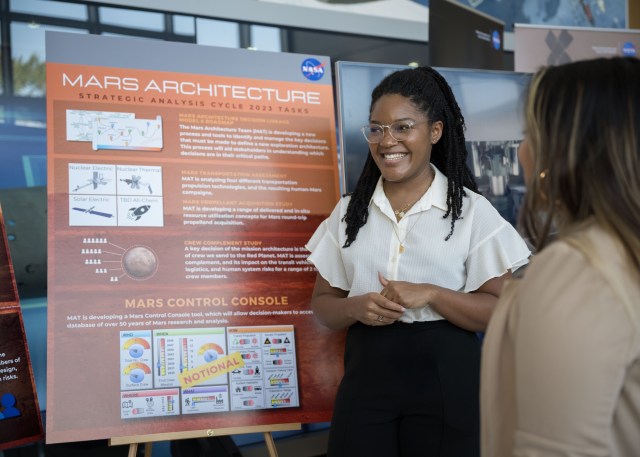 A woman in a lobby smiles in front of a brown poster containing text and images.