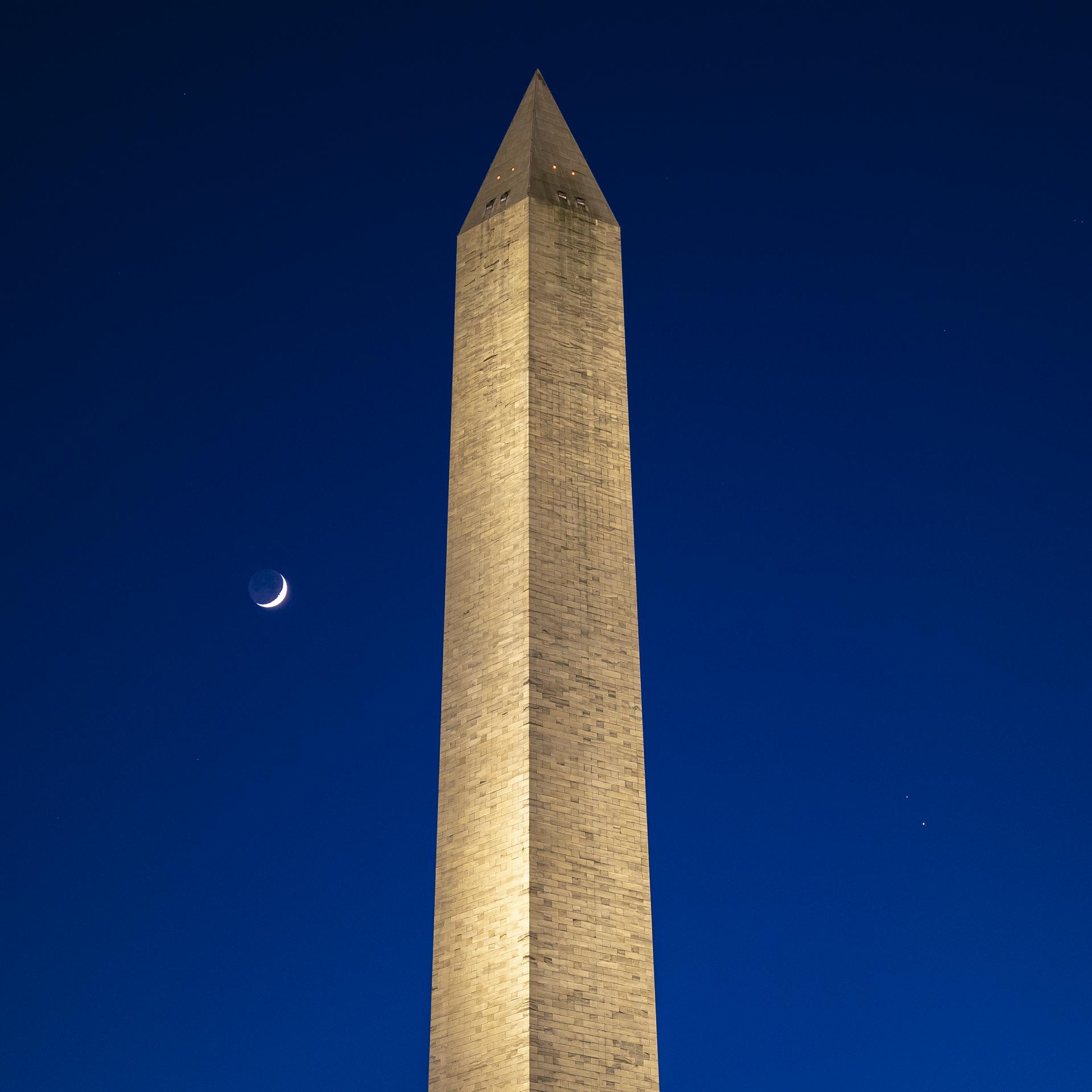 The Washington Monument - a tall, white rectangular monument with a pyramid at the top - is in the center of this image. The Moon (left), Saturn (upper right), and Jupiter (lower right) are in the night sky.