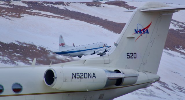 两架飞机在一座被雪和岩石覆盖的山前的图像。前景是一架白色喷气式飞机的尾部，充满了底部和右侧。NASA标志和编号520位于尾部。在这架喷气式飞机的后面，在图像的中间，另一架白色飞机起飞了。它是白色的，有一条蓝色的水平条纹，尾部有NASA的“蠕虫”标志。棕色和白色的山坡填满了画面的其余部分。