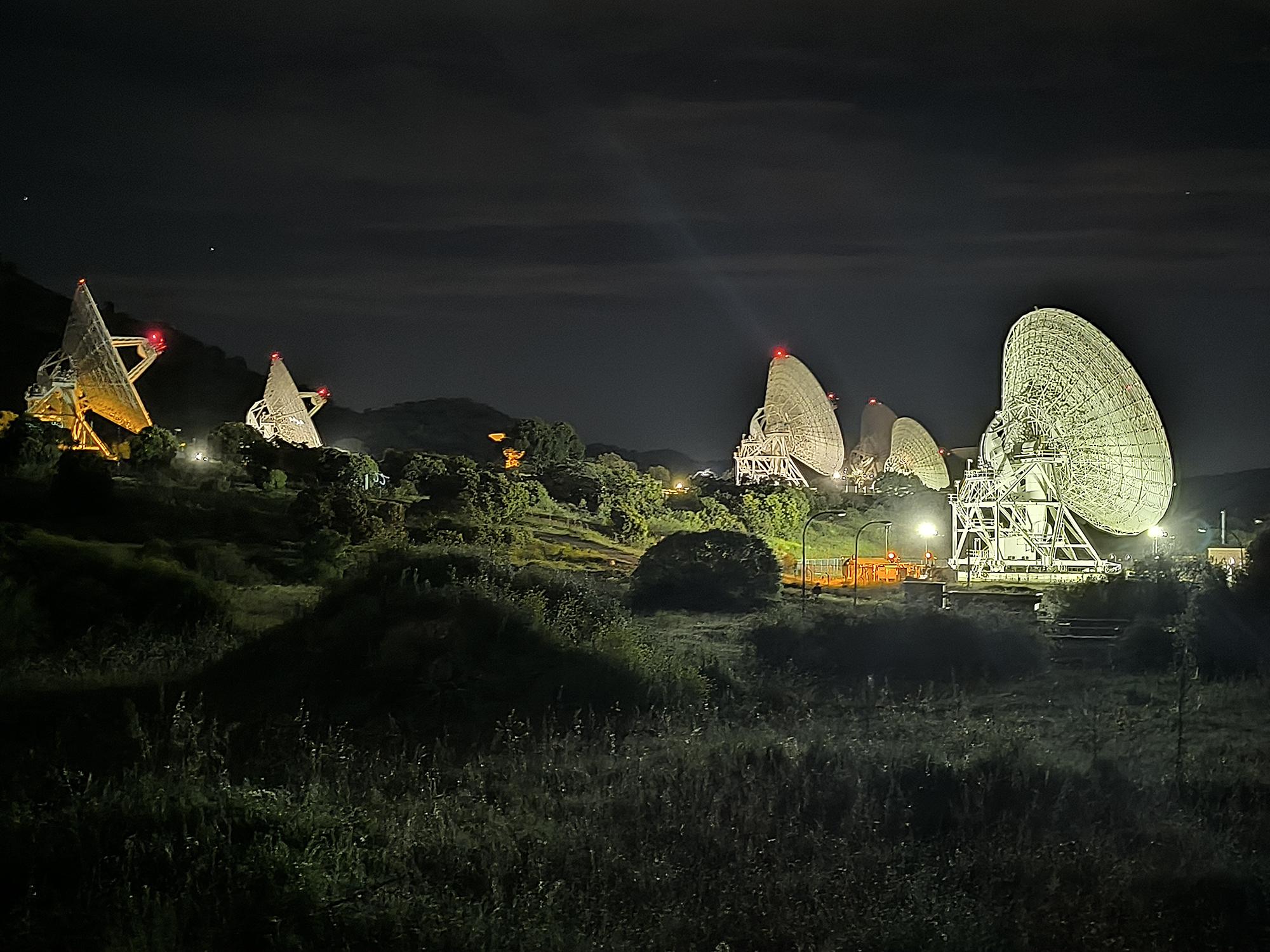 In a nighttime landscape of rolling grasses and trees, 6 large off-white satellites face to the right. Each satellite has bright spotlights near it, but the surrounding area remains mostly dark.