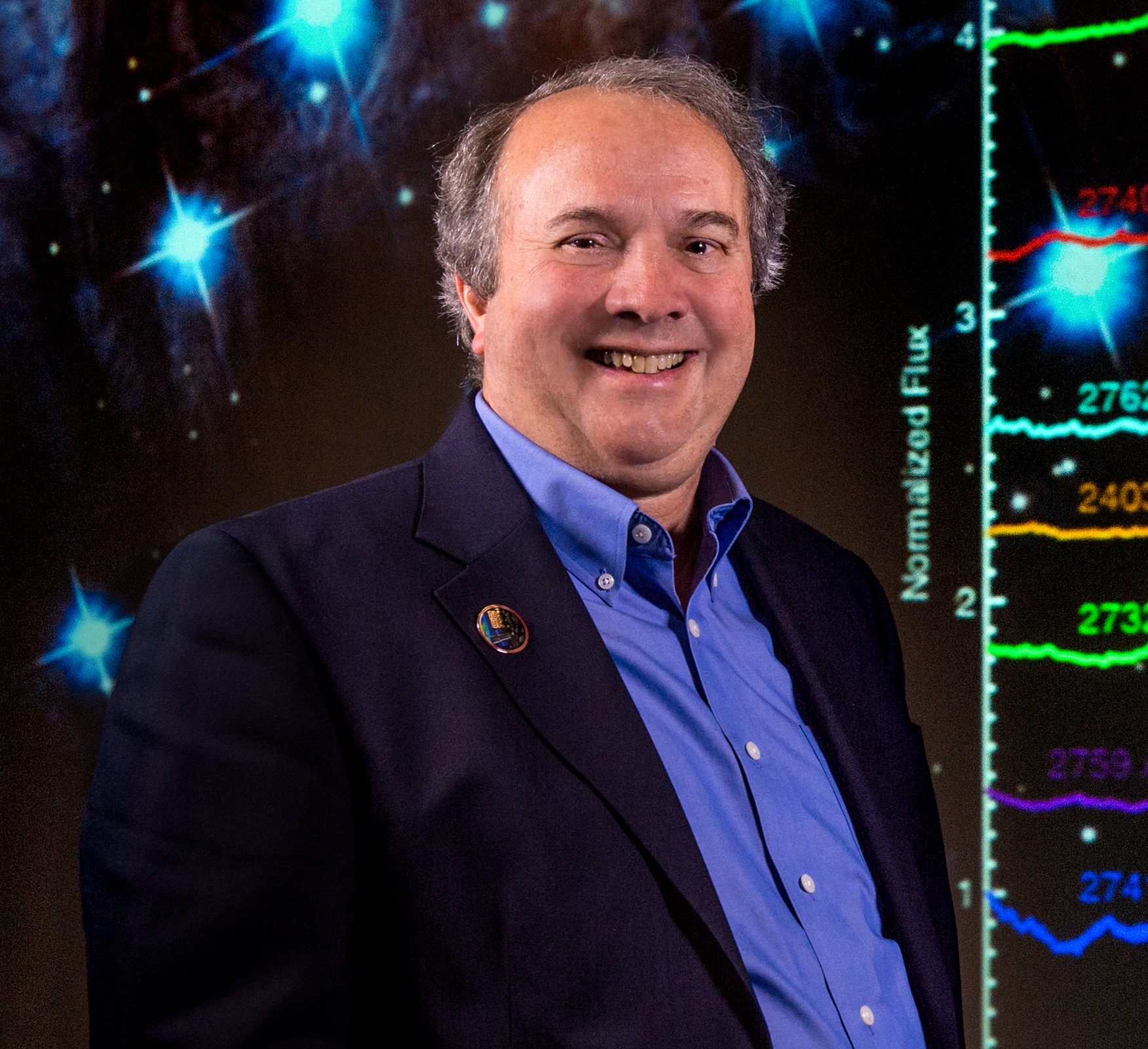 Ken Carpenter smiles wearing a blue dress shirt with a navy jacket. The background is an image of stars with diffraction spikes visible and a chart to the far right.