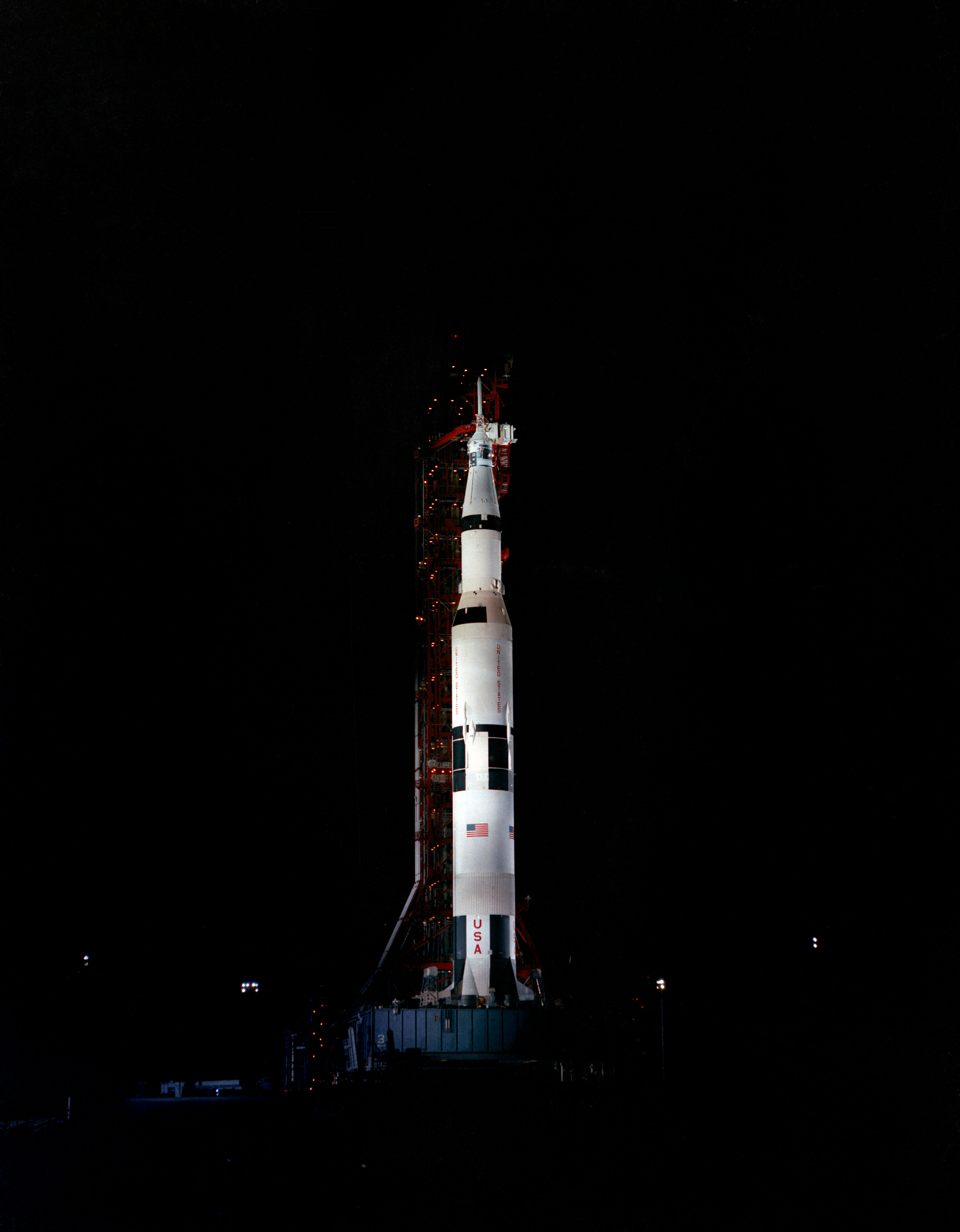 The Apollo 10 spacecraft towers over the launch pad spotlights that light it. It is white, with an American flag and "USA" on the lower portion. It is nighttime.