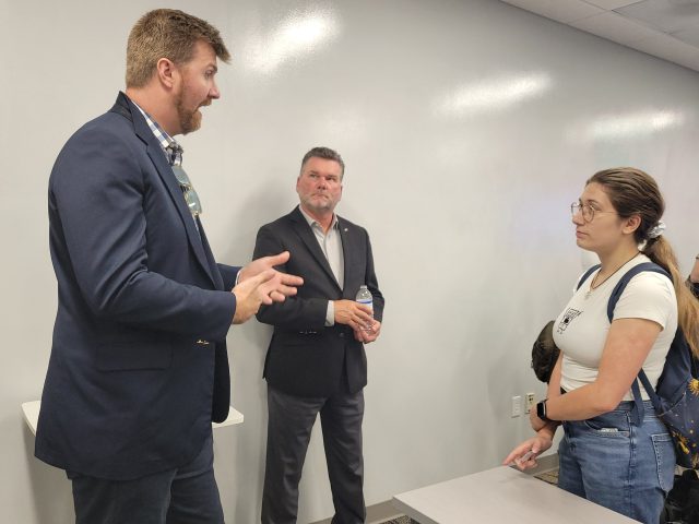 A man in a suit gestures with his hands while talking to a female student as another man stands listening in the background.