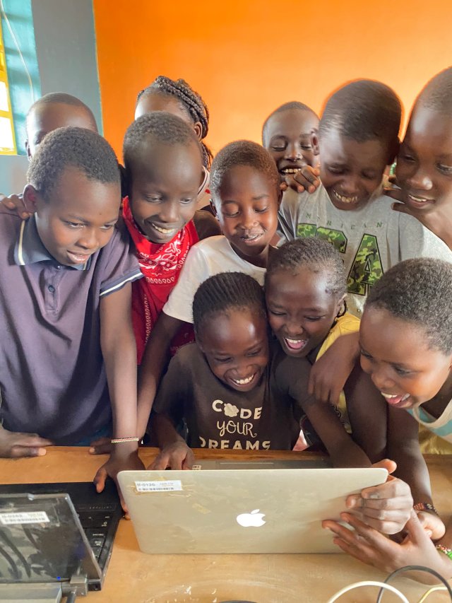 Kenyan students surround a computer laptop. They are smiling and laughing at the screen.