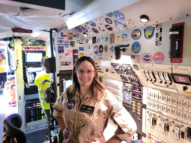 A woman poses, smiling with her hands on her hips, in front of the mission control desk onboard an aircraft. She is wearing a tan flysuit, and the switch board behind her is crowded with buttons, switches, monitors, cords, and stickers.