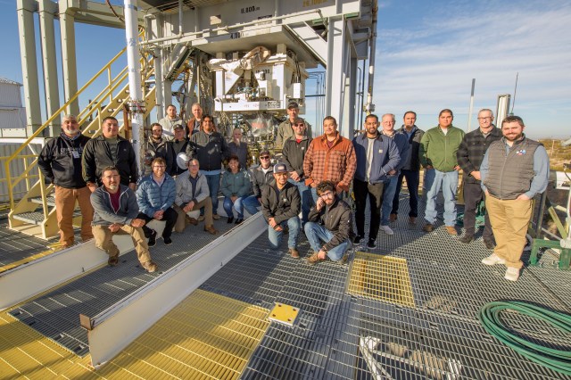 A group of 24 men dressed in casual outdoor clothing stands in front of a white metal test stand used to test a rocket engine injector.