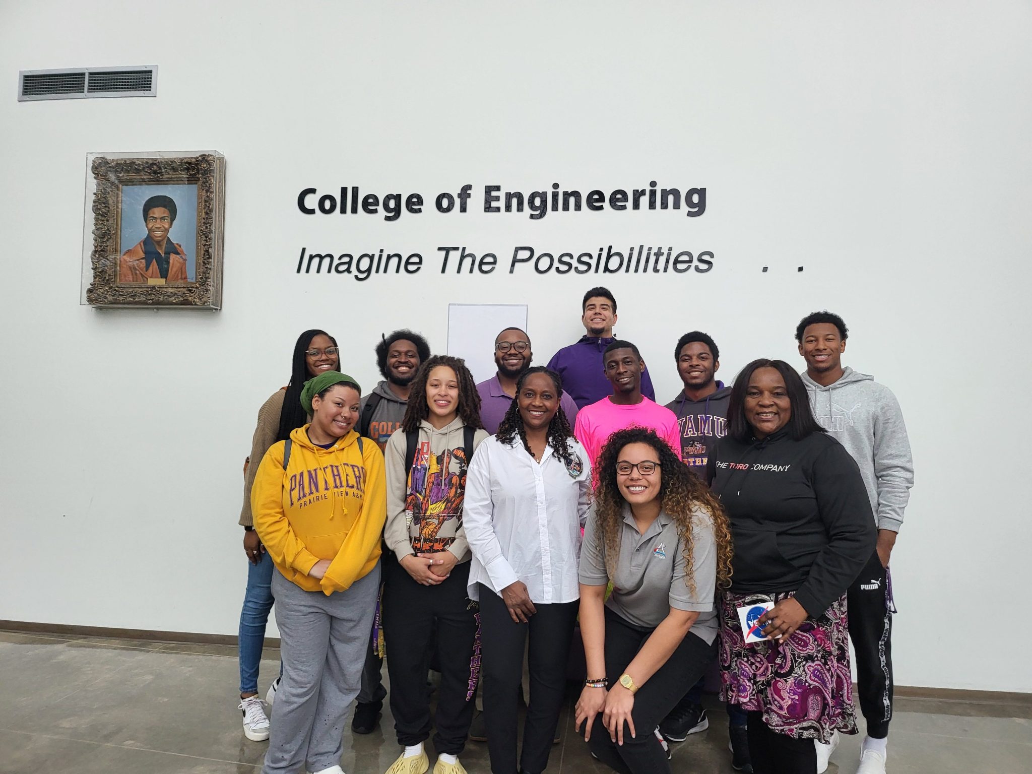 A group of students and faculty members stand in front of a wall that reads "College of Engineering" with the slogan "Imagine The Possibilities." The group is smiling and posing for the photo. A portrait of a man is displayed on the wall to the left of the group.
