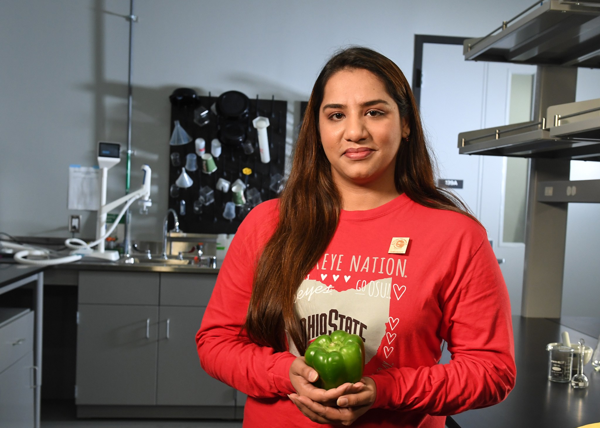 Mehr Un Nisa, a Deep Space Food Challenge Simunaut, stands in a laboratory-like setting wearing a red, long-sleeved Ohio State University shirt. She is holding a green bell pepper.