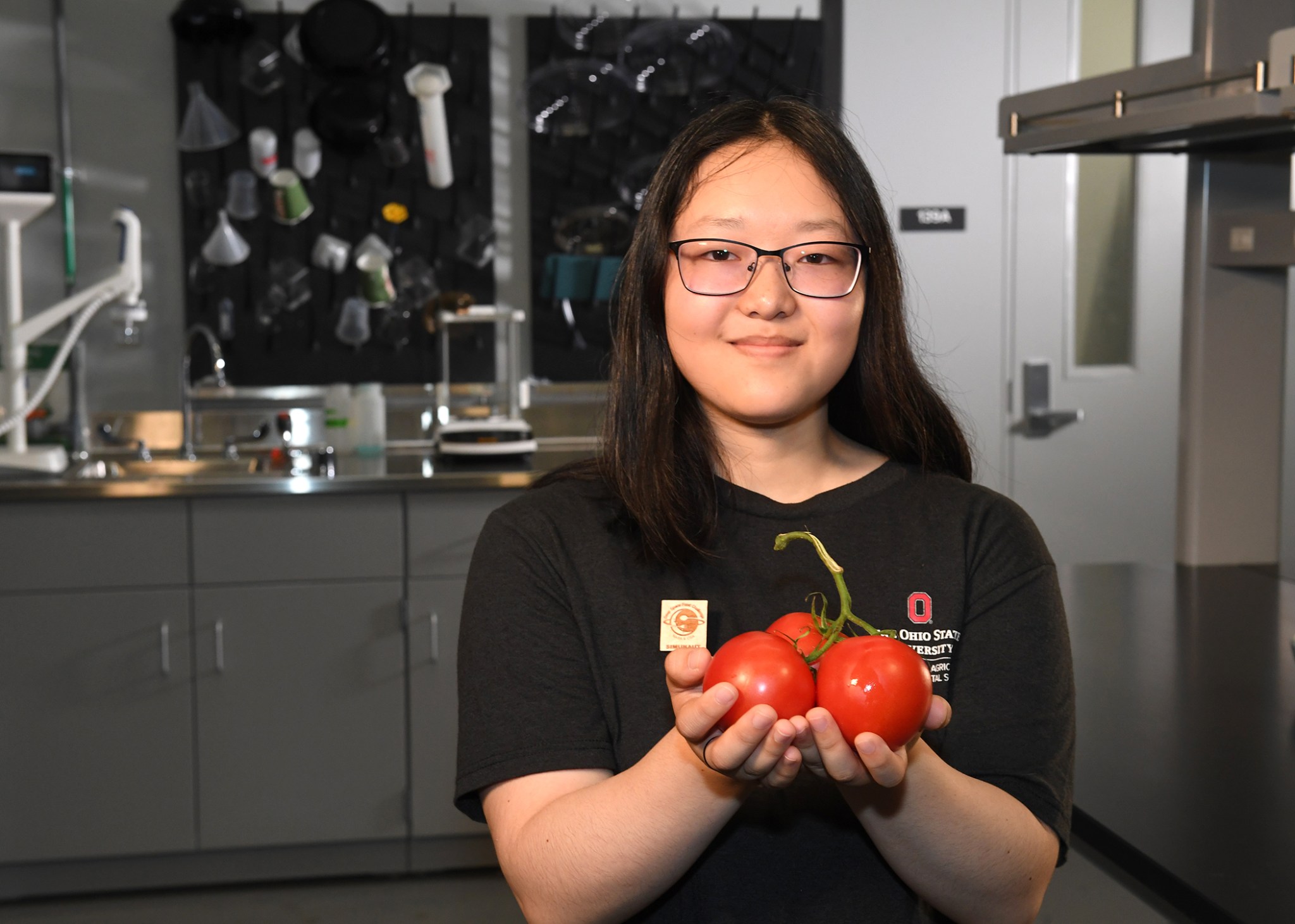 Sakura Sugiyama, a Deep Space Food Challenge Simunaut, stands in a laboratory-like setting wearing a black Ohio State University polo shirt with a gold Deep Space Food Challenge enamel pin. She is holding a bundle of tomatoes on a vine.