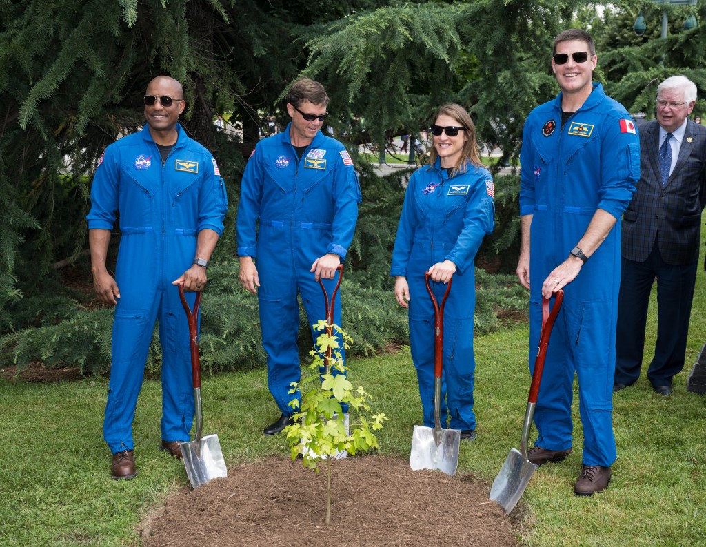 Four astronauts, three men and one woman (second from right), rest their hands on the handles of short shovels that are planted in dirt. They surround a small tree sapling that has bright green leaves.