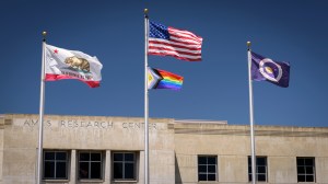 Three silver flagpoles are in front of a building with the words "Ames Research Center" on it. On the left pole is the California state flag. On the middle flagpole are, from top to bottom, the American flag and the Intersex Progress Pride flag. The Intersex Progress Pride flag has a purple circle on a yellow triangle, a five-stripe chevron with colors representing LGBTQI+ people of color and the transgender community, and rainbow stripes. Finally, on the right flagpole, is the NASA flag.