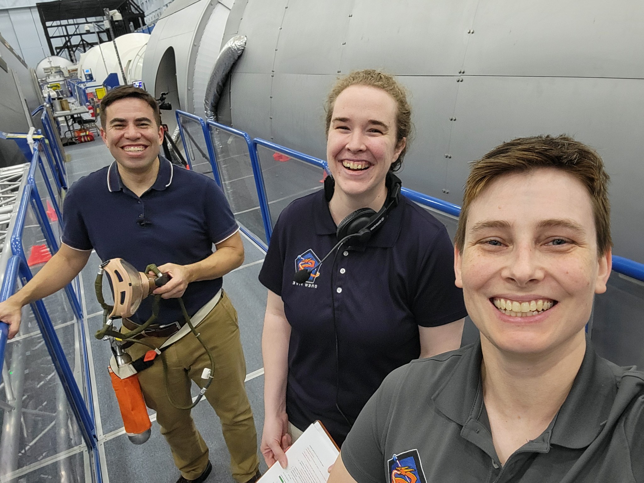 Three people are standing inside a large laboratory environment with metallic structures and equipment in the background.