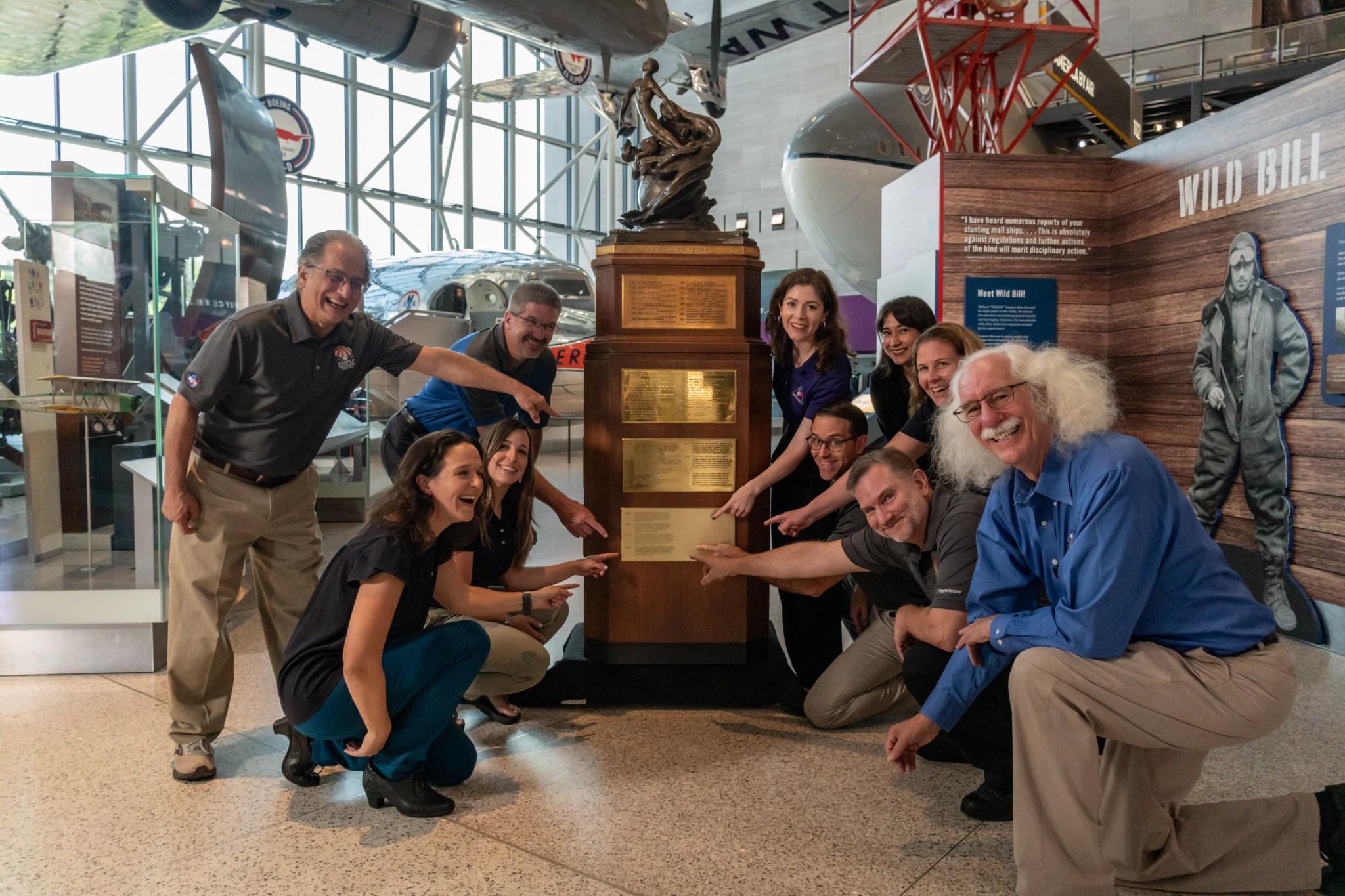 a crowd of people stand around a tall wooden plinth and bronze sculpture; several are smiling and pointing to a plaque on the plinth